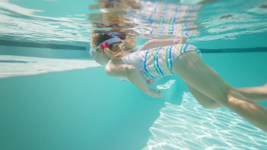 Little girl in striped swimsuit and goggles swimming underwater in a pool