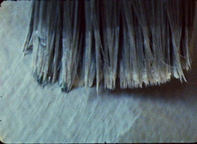 A brush with white bristles at the end of a red handle moves across a textured grey surface