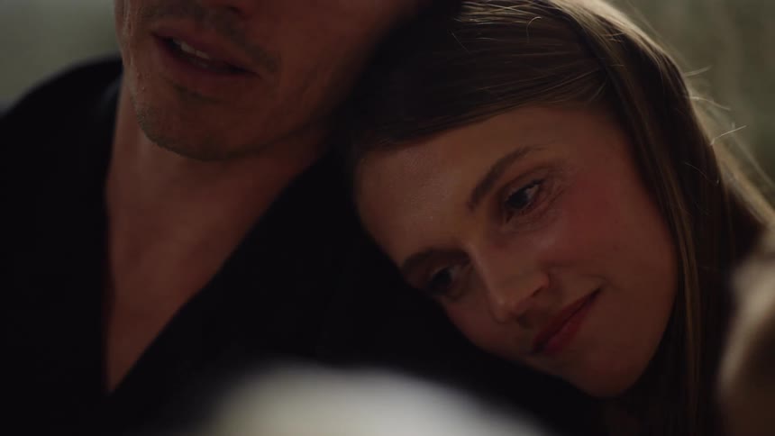 A woman with brown hair rests her head on a mans shoulder while he speaks and smiles down at her as the pair shares an intimate moment