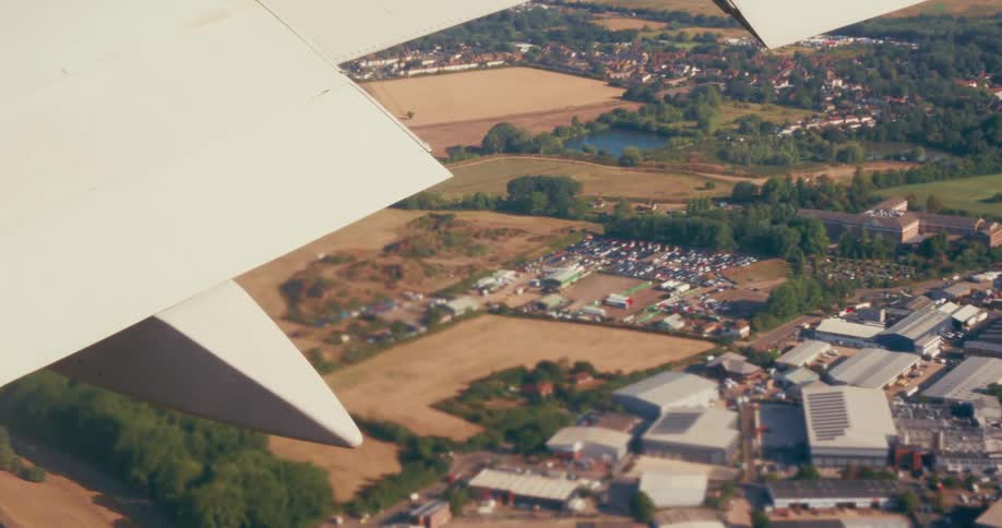 An aerial view from an airplane window shows a sprawling landscape of fields a town industrial buildings and a lake with the planes wing prominently in the foreground