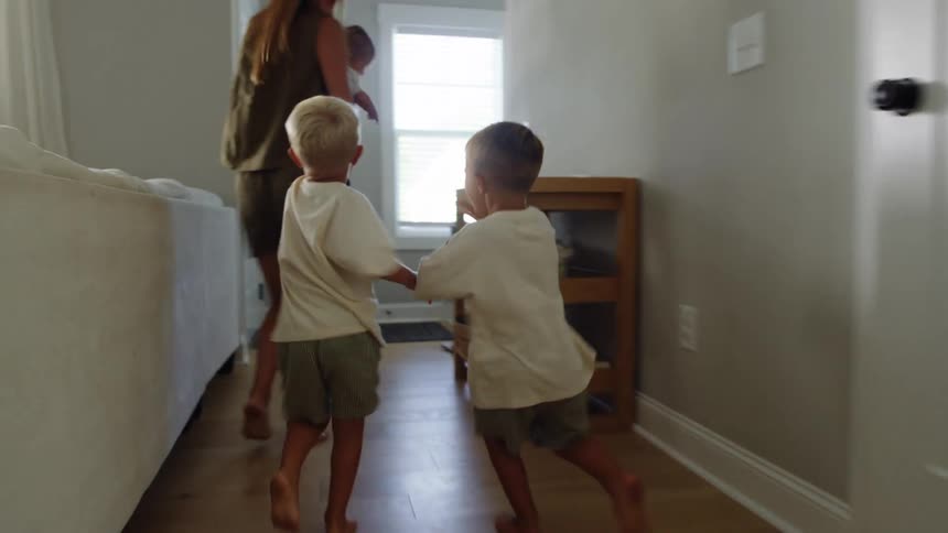 Two young boys playfully chase their mom through the living room as she holds a baby