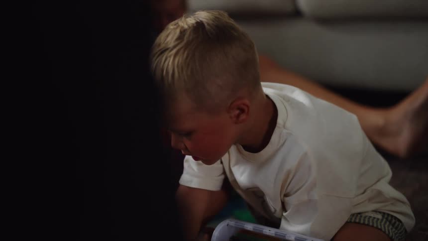 Young blonde boy playing with toys in a living room setting and theres a person holding a baby in the background