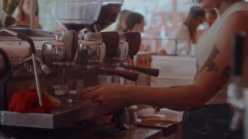 The video shows a closeup shot of a cafe barista as they prepare a coffee drink using an espresso machine the barista is seen from the chest up and their hands are visible as they work the machines components including a portafilter and tamper