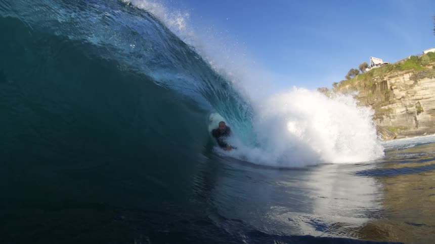 A surfer tucks into a curling wave near rocky cliffs under clear skies