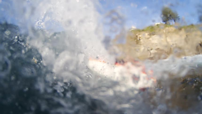 A close view shows a man with a beard and a black wetsuit on a boogie board riding a crashing wave that moves toward a light colored cliff with green plants on top