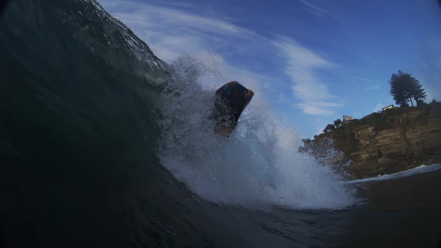 The video shows an underwater view of a surfer and a wave the sky is visible above the water with a cliff face in the background