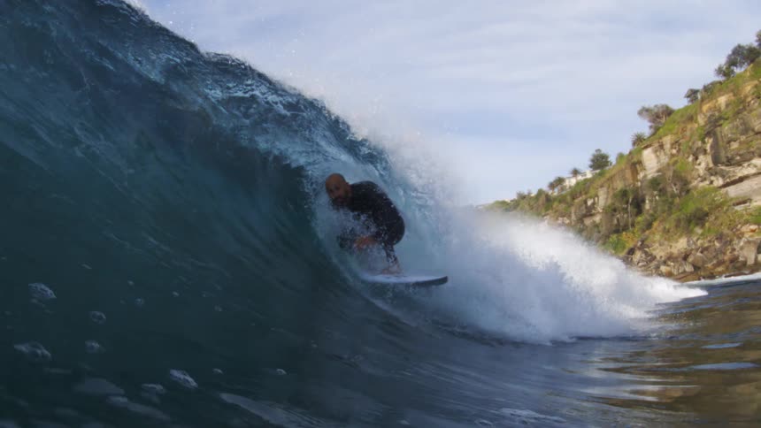 Surfer rides inside the barrel of a blue wave near the rocky shore