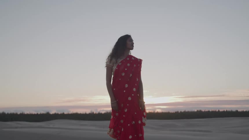 A hindi woman in a red sari with gold embroidered designs stands on a sandy beach at sunset with her her head turned to the right