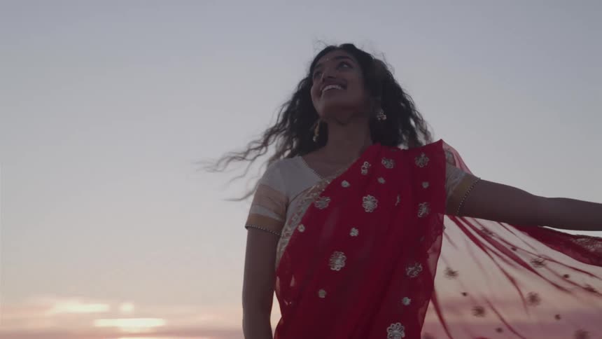 A young hindi woman wearing a red sari and jewelry on her arms is dancing on a beach at sunset with her hair and the long fabric of her sari blowing in the wind