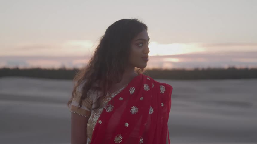 A young woman wearing a red sari stands outside at sunset