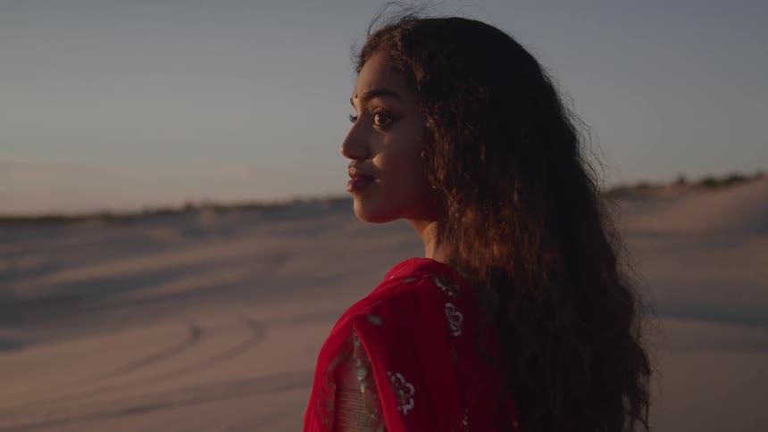 A young woman wearing a red sari stands on the desert sand at sunset