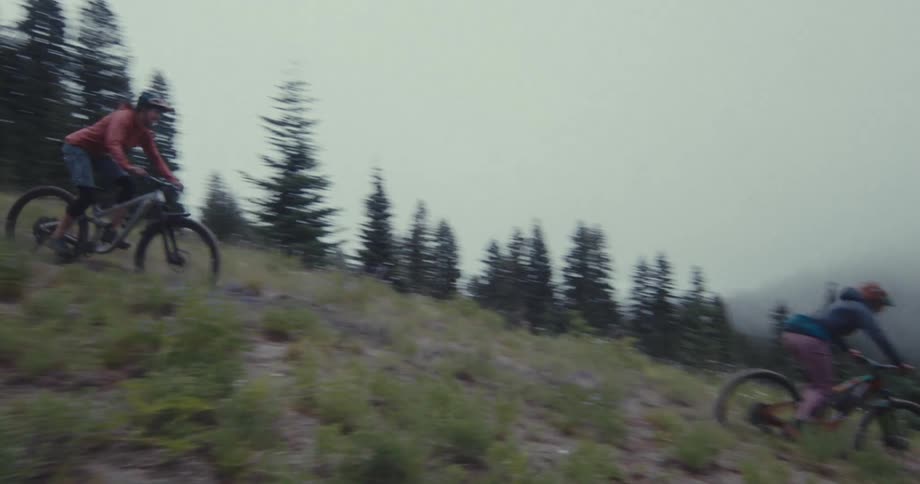 Two mountain bikers ride down a steep grassy hill lined with evergreen trees under a cloudy sky the bikers who appear to be a man and a woman speed past the camera kicking up dirt as they go