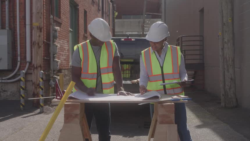 A pair of coworkers wearing white hard hats and yellow safety vests are examining blueprints on a wooden table in a sunny alley