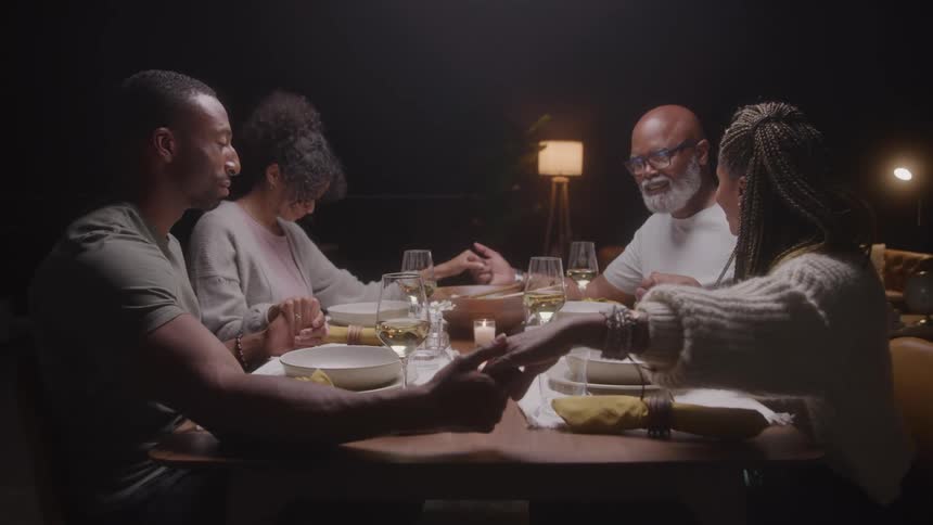 A family of four is seated at a dimly lit dinner table holding hands bowing their heads and praying over their meal