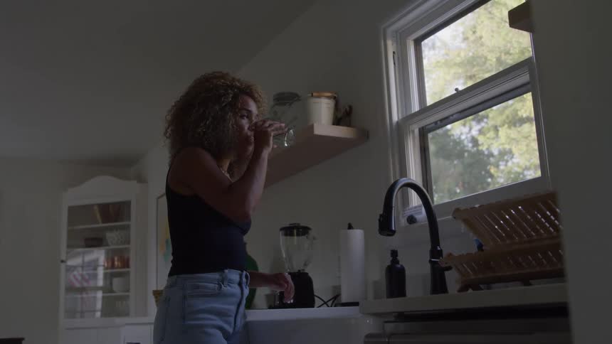 A woman smiles while standing in her kitchen drinking a glass of water