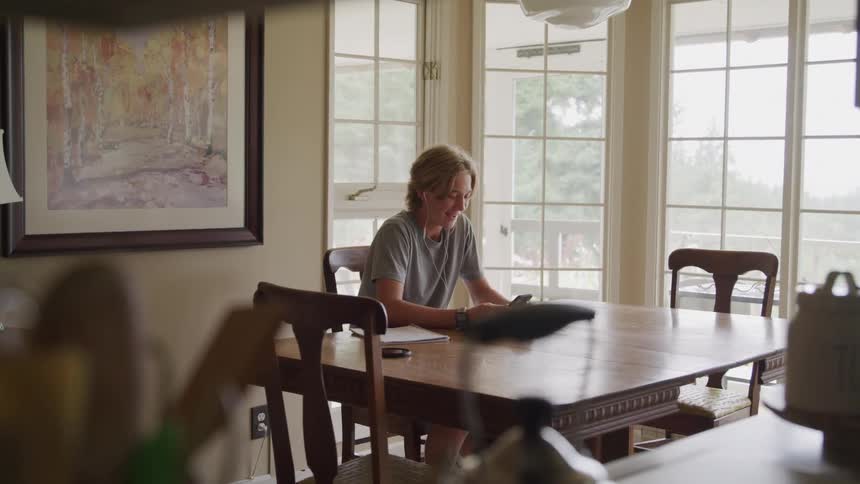 A smiling person with light colored hair is sitting at a wooden dining table using a cellphone and talking