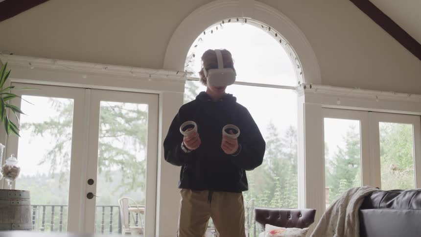 A male teenager playing a virtual reality game while standing in a living room during daytime