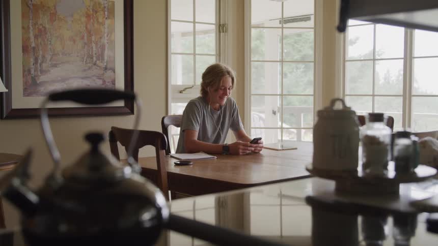 The video shows a person with a blond ponytail sitting at a dining table looking at their phone they are smiling and appear relaxed with a notebook pen and tablet also on the table