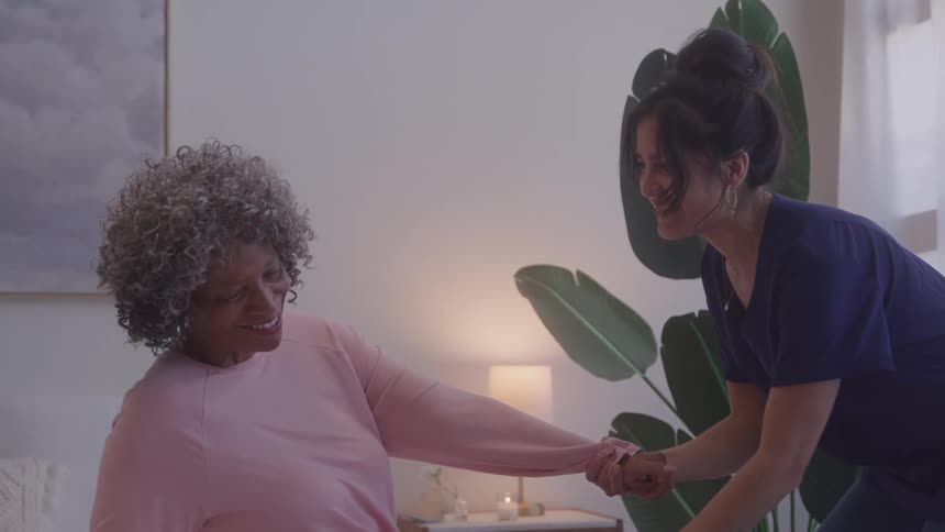 A happy female patient with gray hair and a pink shirt is smiling and sitting up in her bed while a female medical staff member wearing dark blue scrubs holds her hands