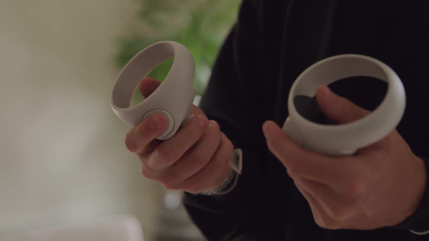 A close view of a persons hands shows them holding two white virtual reality controllers with circular frames