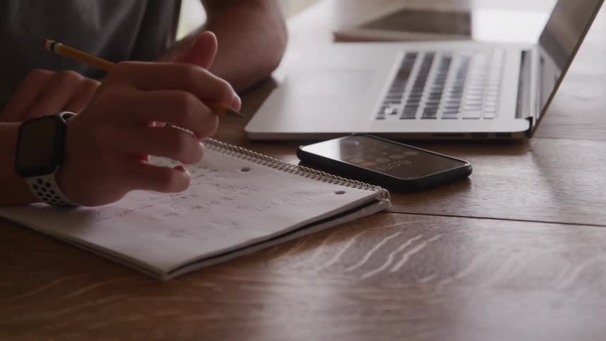 A person wearing a smartwatch writes in a notebook while checking a phone beside a laptop