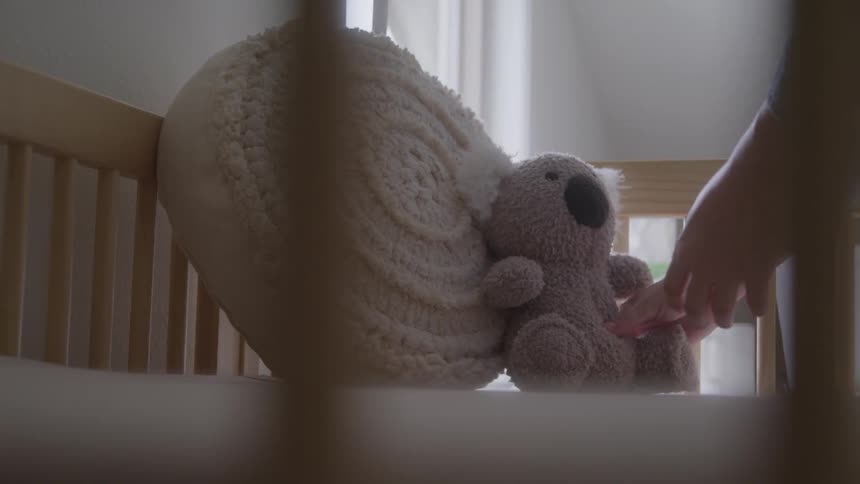 A hand places a textured beige pillow and a small stuffed grey koala into a wooden crib