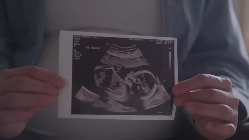 A pregnant woman holds a sonogram printout of two fetuses in the foreground as she looks down at the image with a smile