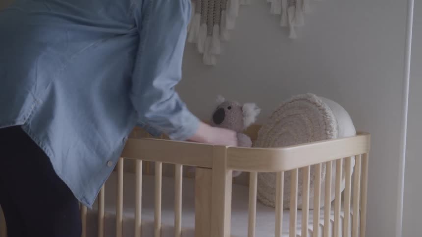 A woman walks into a home nursery to place a pillow in a wooden crib