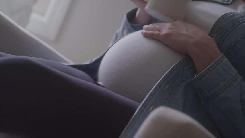 Hand resting on the belly of a pregnant woman holding a mug