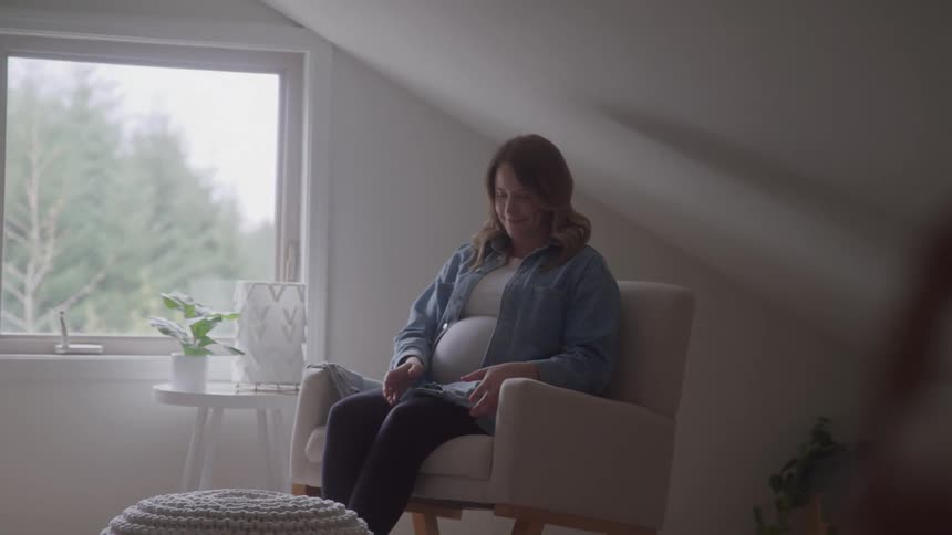 A pregnant woman with brown hair sits in a light colored armchair and holds up a small blue garment smiling as she looks down and places it on her large belly