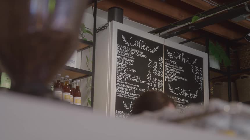 A chalkboard menu lists various coffee and drink options inside a caf as a person stands near the menu