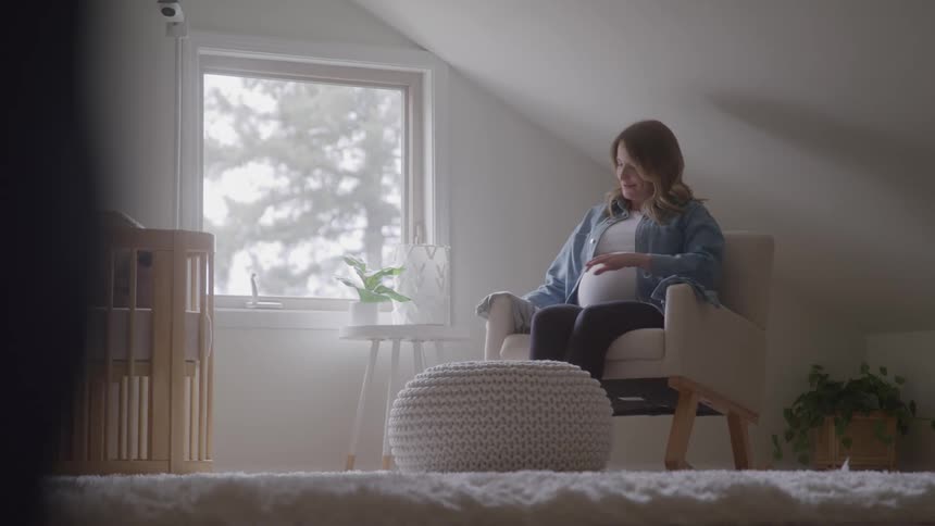 The video shows a pregnant woman sitting in a cozy room looking at baby clothes she is holding a small blue outfit and smiling as she examines it012describe 2 lines