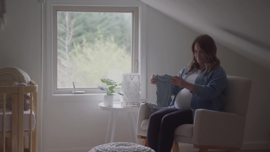 The video shows a pregnant woman sitting in a rocking chair in a babys nursery she is holding up a small blue baby garment smiling at it as she examines the details