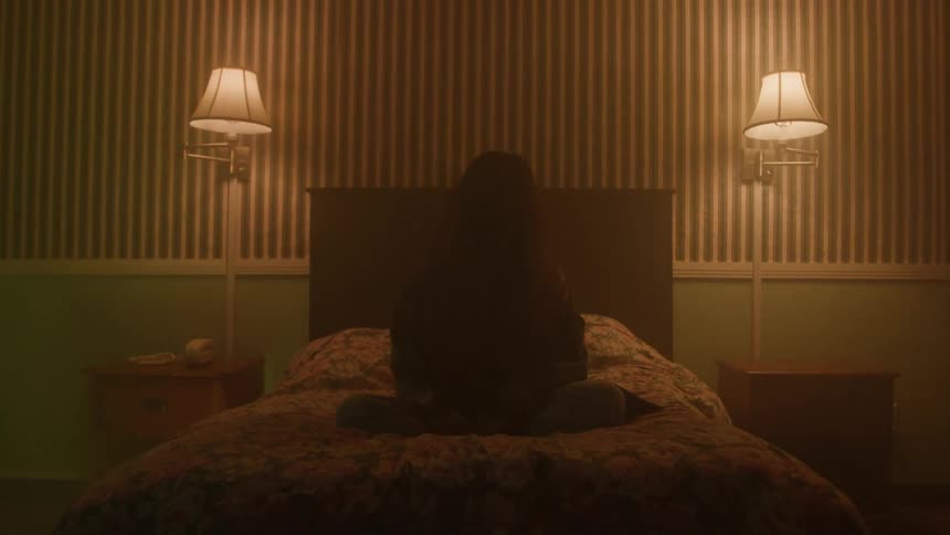 A woman sits on a bed in a dimly lit room with two lamps casting soft light on either side