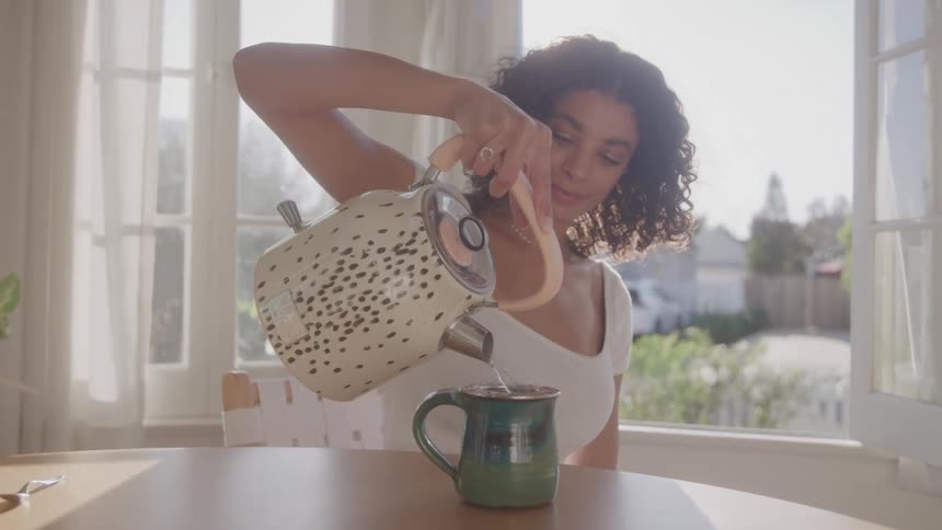 A woman with dark curly hair is sitting at a wooden dining table and pouring a hot beverage from a white and black kettle into a green ceramic mug