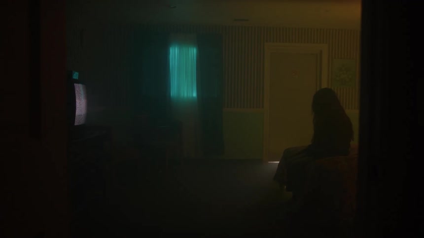 A silhouetted figure sits on a bed facing a staticfilled tv teal window light cutting the dark