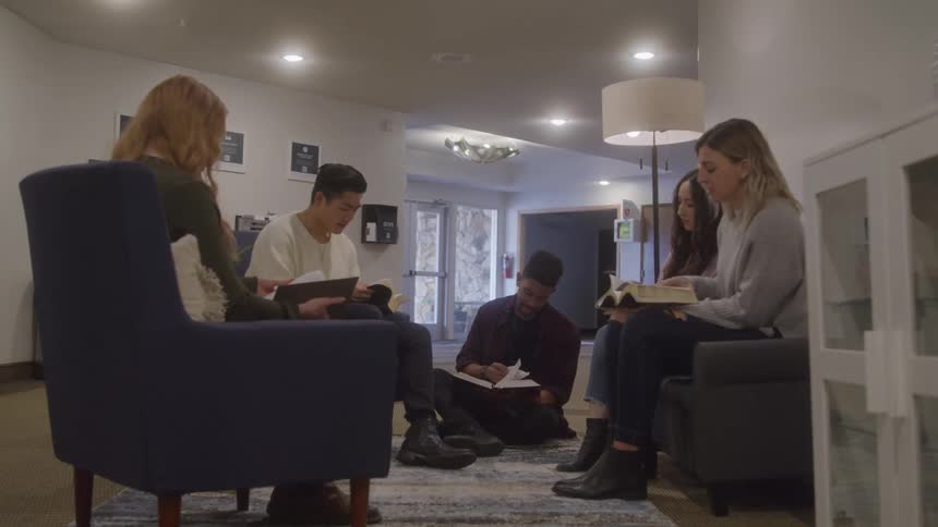 A group of five friends including a man sitting on the floor and four other people in two armchairs are sitting in a circle and talking while holding notebooks and books in a well lit room
