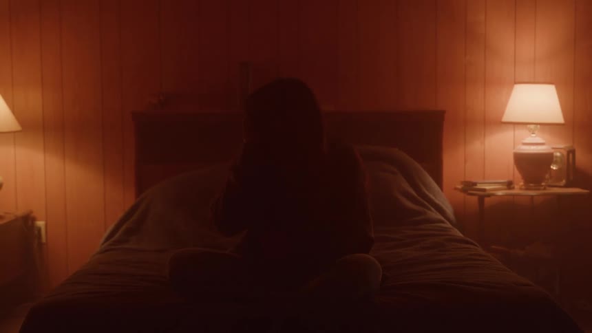 Someone sits on a bed in warm lamplight the room glowing with moody amber tones