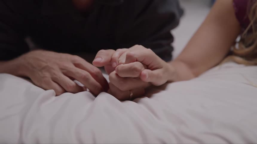 Two people lying down holding hands on the bed