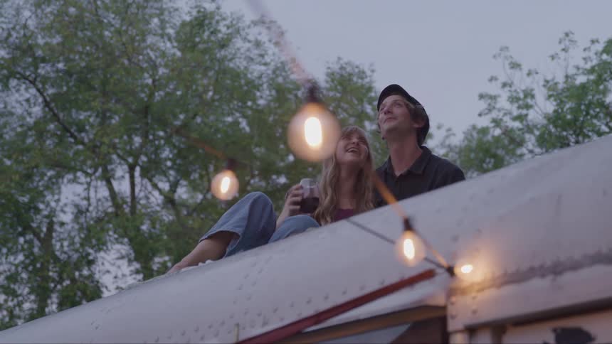 A young couple relaxes on a bus roof under warm string lights at dusk smiling and chatting