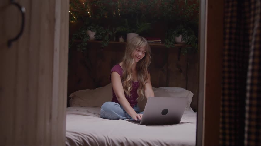 A person sits on a bed with a computer on their lap and a bookshelf with plants behind them