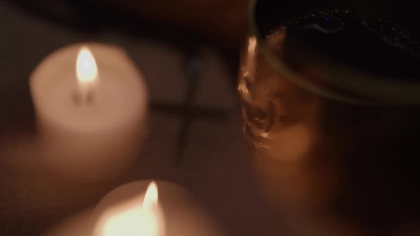 Two burning candles are near a metal cup full of a dark liquid