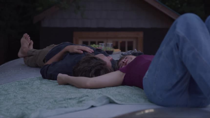 A woman in jeans and a man with bare feet are lying on a patterned blanket on what appears to be the top of a vehicle looking up at the sky together in the evening
