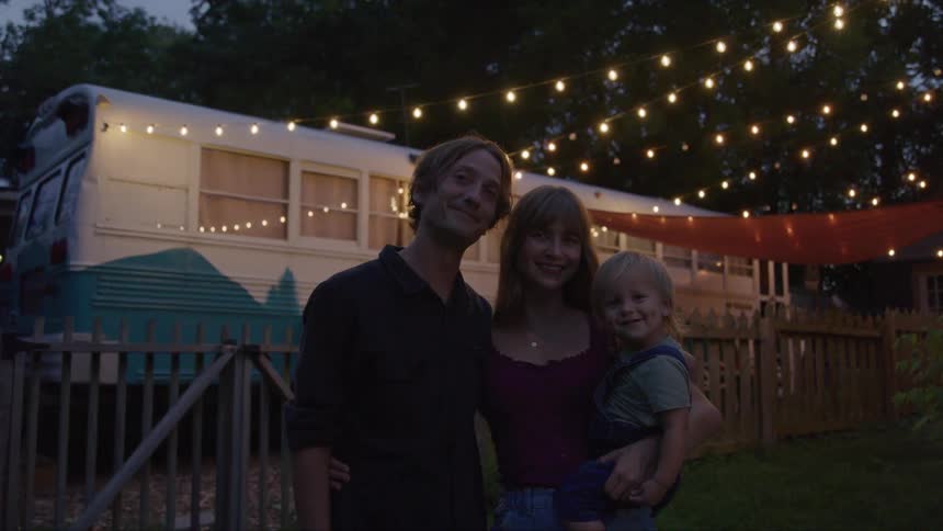 A couple and their toddler pose at dusk beneath patio string lights a converted bus and picket fence framing their cozy yard