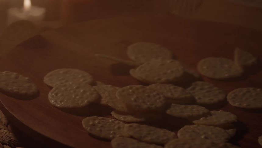 The video shows a hand dropping what appears to be tortilla chips or food pieces onto a round wooden tray a blurry lit candle is visible in the background