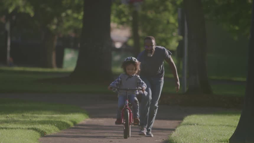 The video shows a father and his young child on a sidewalk in a parklike setting the child wearing a helmet is riding a small red bicycle while the father runs alongside helping to balance the bike