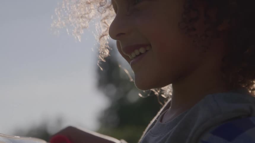 A girl with curly hair smiles while blowing bubbles with a wand