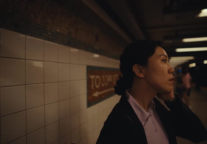 A woman in a subway station standing close to the back wall covered in white tiles looks nervous as she wipes sweat from her face with her hand