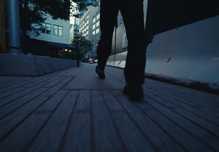 The video shows a person wearing a dark coat and pants walking on a sidewalk at night the streetlights illuminate the path and a building with a mural is in the background