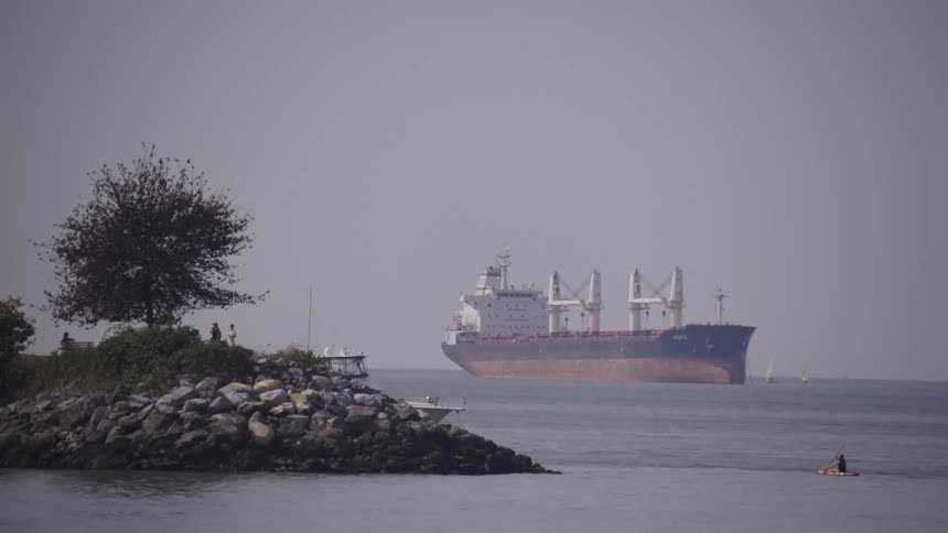 The video shows a cargo ship in the water with a rocky shoreline and a small sailboat in the distance the sky is smoke filled and the overall color palette is muted gray and blue